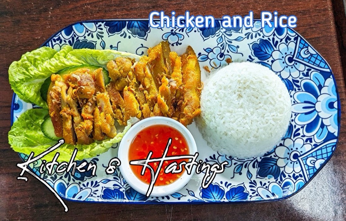 A blue-and-white plate of grilled chicken pieces on lettuce with steamed rice and a pot of sweet chilli sauce; the photo has the words Chicken and Rice written on it