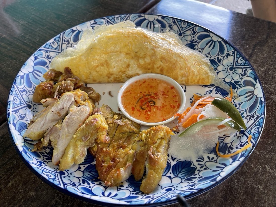 A blue-and-white plate with a folded golden omelette, pieces of grilled chicken and a small dish of dipping sauce