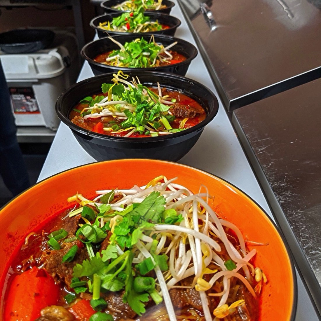 A bright orange bowl of noodle soup topped with bean sprouts, spring onion and herbs, with more bowls lined up behind it