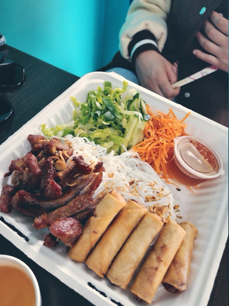 A takeaway tray of vermicelli noodles with grilled pork, spring rolls, cucumber, carrot and a pot of dipping sauce