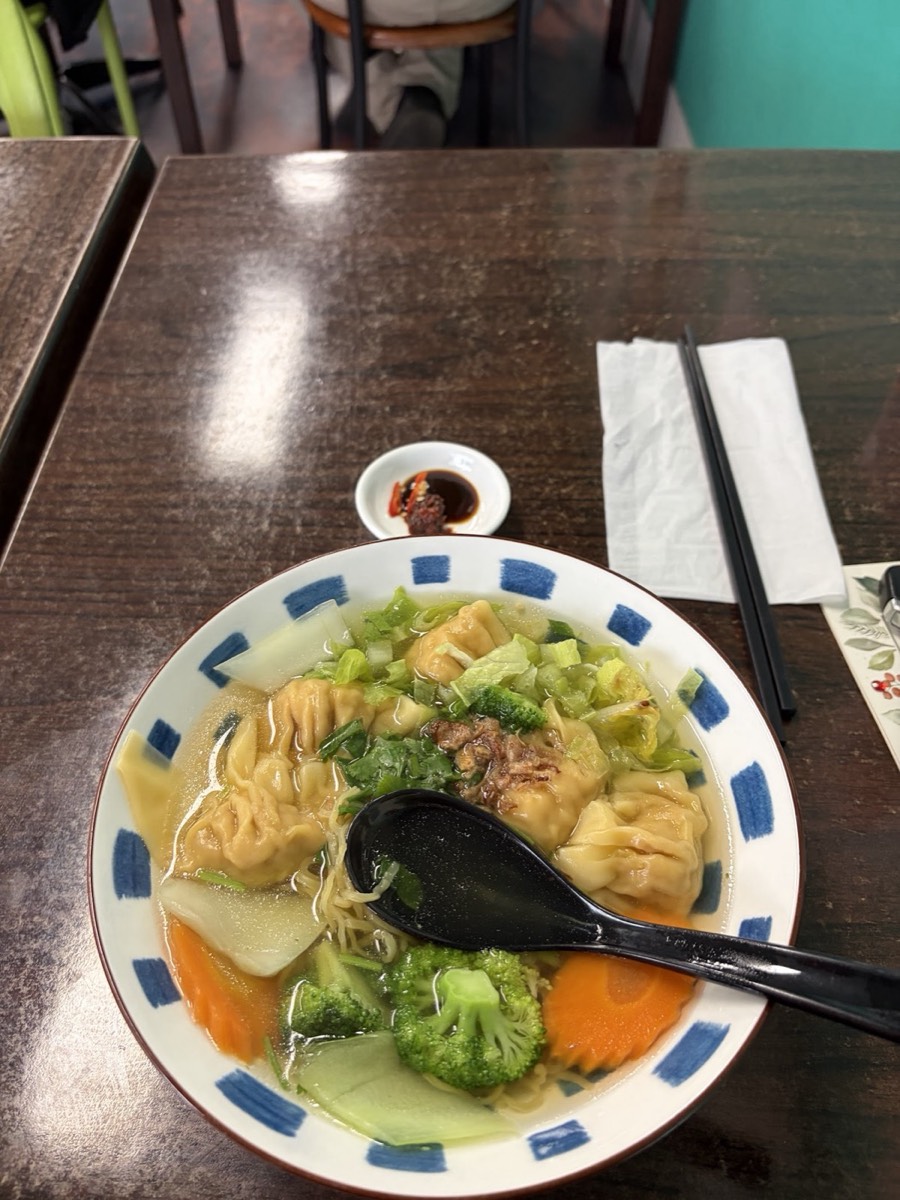 A blue-and-white bowl of wonton noodle soup with broccoli and carrot, a black spoon and chopsticks on a dark table